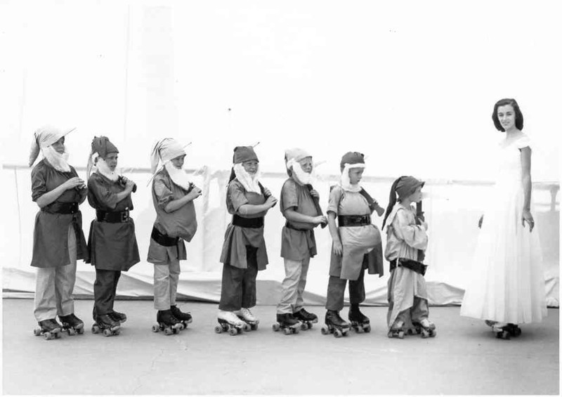 Black-and-white photo: six children in caps and aprons stand in a line wearing roller skates; an adult woman stands at the right.